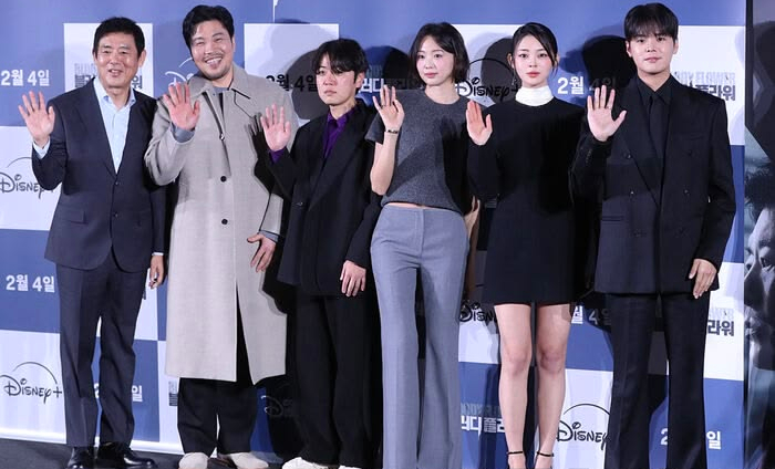 A group of six individuals, dressed in formal and stylish attire, stand on a red carpet against a Disney+ backdrop, smiling and waving at the camera.