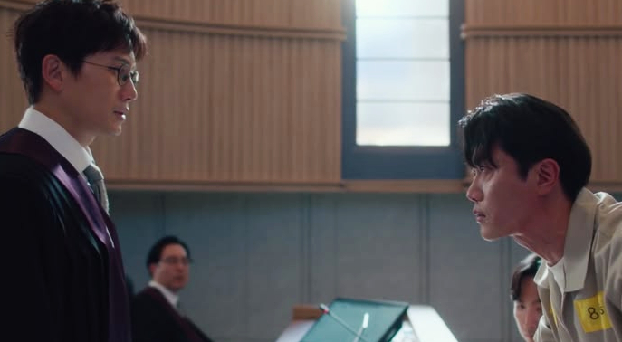 A tense courtroom scene with a lawyer in a suit and a man in a prison uniform standing confrontation-ally. A window casts light, creating a dramatic atmosphere.