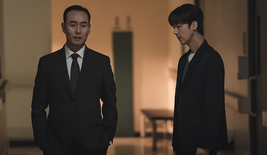 Two men in formal suits stand in a dimly lit corridor. One man appears serious, while the other looks pensive. The setting conveys a tense atmosphere.