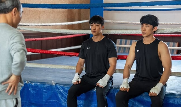 Two boxers in black tank tops and hand wraps sit attentively on a boxing ring edge, facing an older man. The atmosphere is focused and intense.
