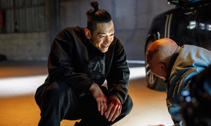 A man with a topknot, wearing dark clothes, crouches with blood on his hands, smiling at another man kneeling. The scene is tense and dramatic.