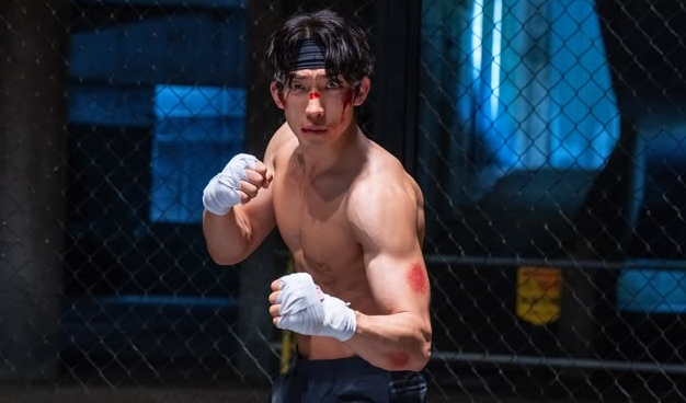 A shirtless man with boxing tape on his hands stands in a fighting stance inside a dimly lit cage. His intense gaze and muscular build convey determination.