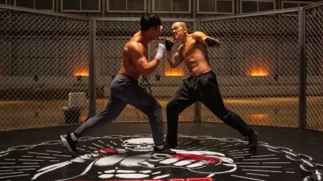 Two shirtless fighters spar intensely in a chain-link cage, surrounded by a dimly lit arena. The atmosphere is tense and action-packed.