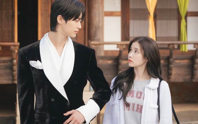 A tall man in a black suit gazes at a woman in casual attire. They stand closely, exchanging a serious yet affectionate look in a traditional setting.