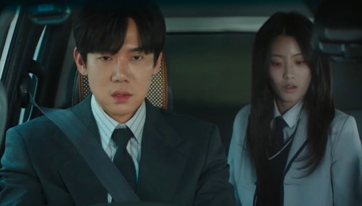 A man in a suit drives a car, looking serious. A woman in a lab coat sits in the back seat, appearing concerned. The scene is tense and focused.