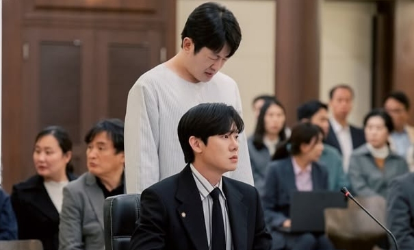 A serious courtroom scene with a man in a suit sitting at a desk and another in casual clothes standing behind him, surrounded by attentive people.