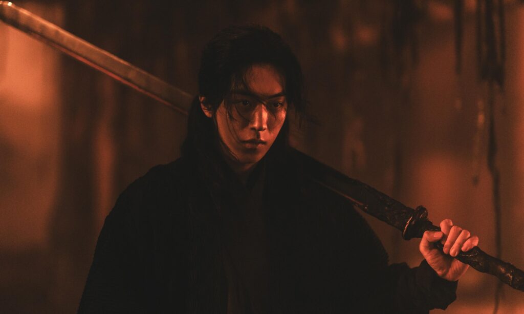Young warrior with long dark hair holds a large sword over his shoulder in a dimly lit, moody environment.