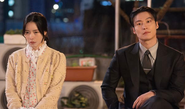 A man in a suit and a woman in a cardigan sit side by side outdoors at night, both looking towards the camera with neutral expressions.