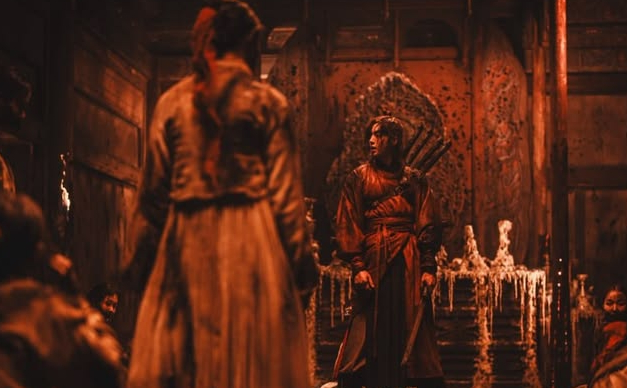 A person in red traditional clothing stands in a dimly lit, ornate room facing another figure; melted candles and intricate wall carvings are visible in the background.