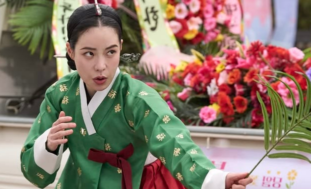 An actress in traditional Korean hanbok, wearing a green jeogori with gold embroidery and a red skirt, holds a small palm frond while performing outdoors near a display of colorful flowers.