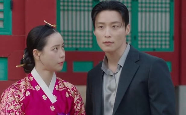 A woman in a vibrant pink hanbok with gold embroidery looks up at a man in a modern black suit and grey shirt against a backdrop of traditional Korean architecture.