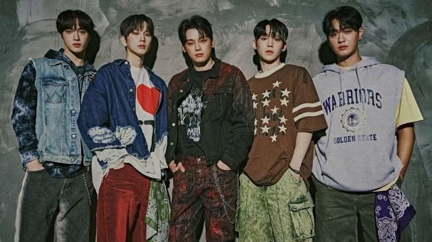 Five young men stand in a row against a textured grey wall, wearing casual, layered street-wear including denim, graphic tees, and a Warriors hoodie.
