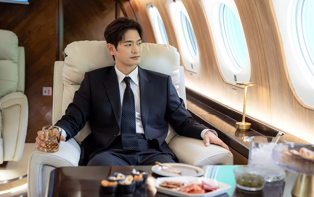 A man in a dark suit and tie sits in a white leather seat inside a luxury airplane cabin, holding a glass of amber liquid while looking away.