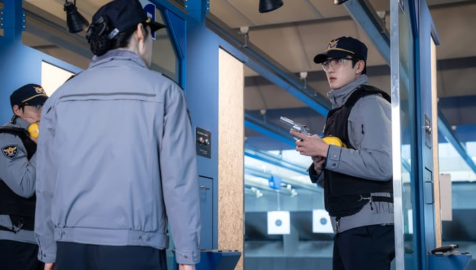 Two police officers in grey uniforms and caps stand in a blue shooting range, one holding a silver revolver while wearing yellow ear protection gear.