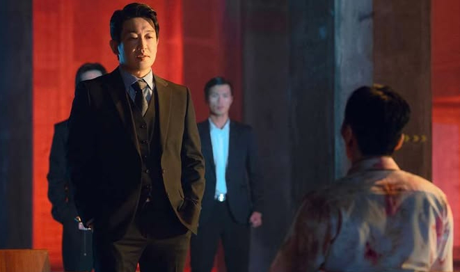 A man in a dark suit and vest stands before a person in a blood-stained shirt, with two other men in suits standing in the dimly lit red background.
