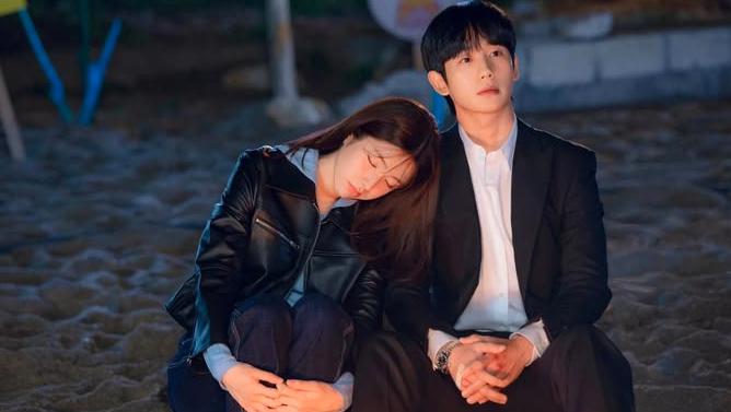 A young woman in a black leather jacket rests her head on the shoulder of a man wearing a black suit and white shirt while sitting on the sand.