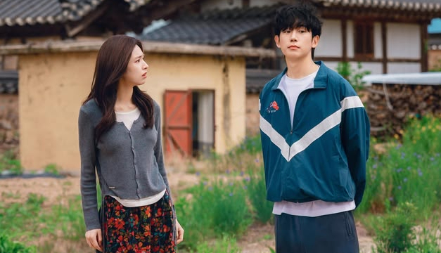 A woman in a grey cardigan and floral skirt looks at a man wearing a teal and white track jacket standing in a rural area with traditional houses.